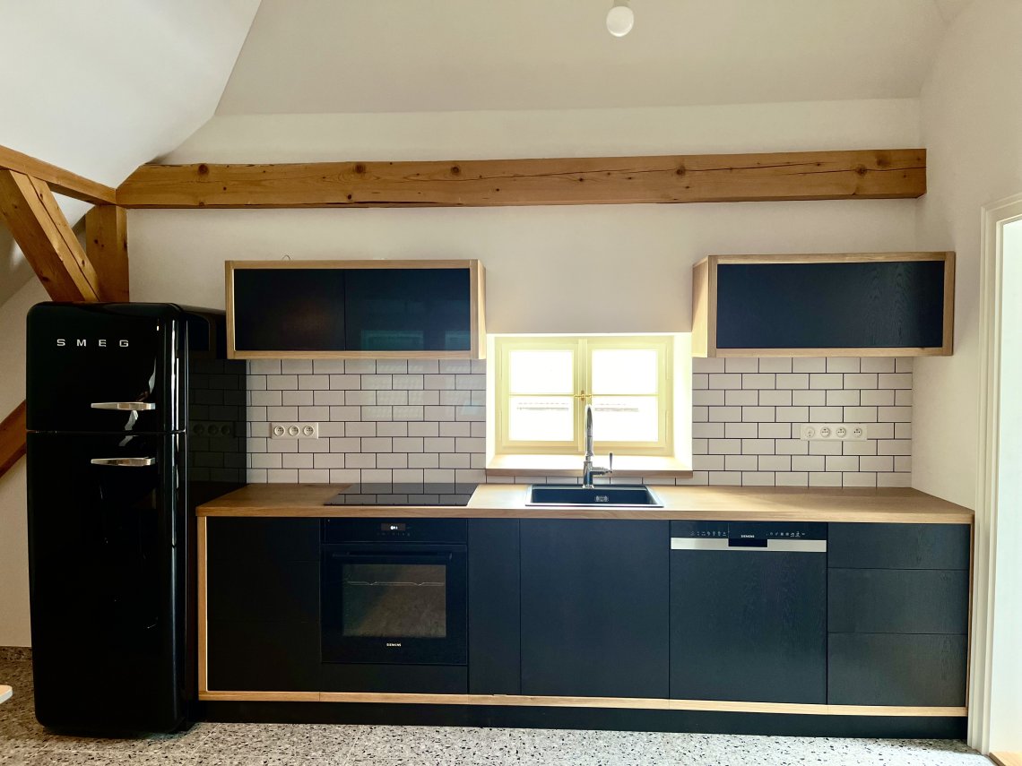 Rovná kuchyňská linka dlouhá 5,2 metru s masivní dubovou pracovní deskou o síle 38 mm, ošetřenou voskovým olejem OSMO pro zátěžové plochy a kontakt s potravinami. Dvířka a čela zásuvek dýhovaná LTD dub, ošetřena barvou Tikkurila - symphony - antracit. Výklopný systém horních dvířek Blum Aventos, který je společně se zásuvkami řešen bezúchytkovým systémem otevírání ''push to open''. Díky tomu bylo možné zachovat kompaktní linie a plochy, které jsou pro tento design charakteristické.